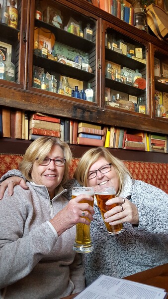 two women sitting touching glasses of beer together. Cheers. Crawl2Walk coming in 2021